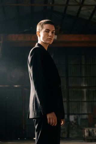 Person in a dark tailored suit standing in an industrial warehouse, glancing back over their shoulder with backlight streaming through the space