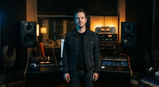 Music producer standing in a dimly lit recording studio surrounded by mixing consoles and studio monitors